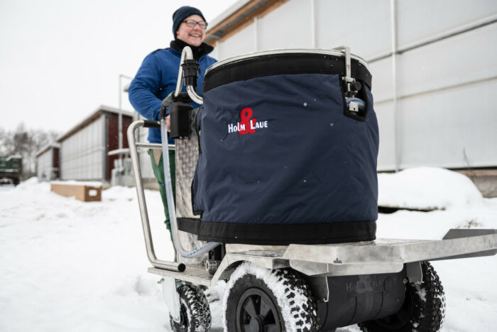 Auf diesem Foto ist ein MilchTaxi mit Mantel zu sehen. Es wird von einer Landwirtin durch den Schnee geschoben.